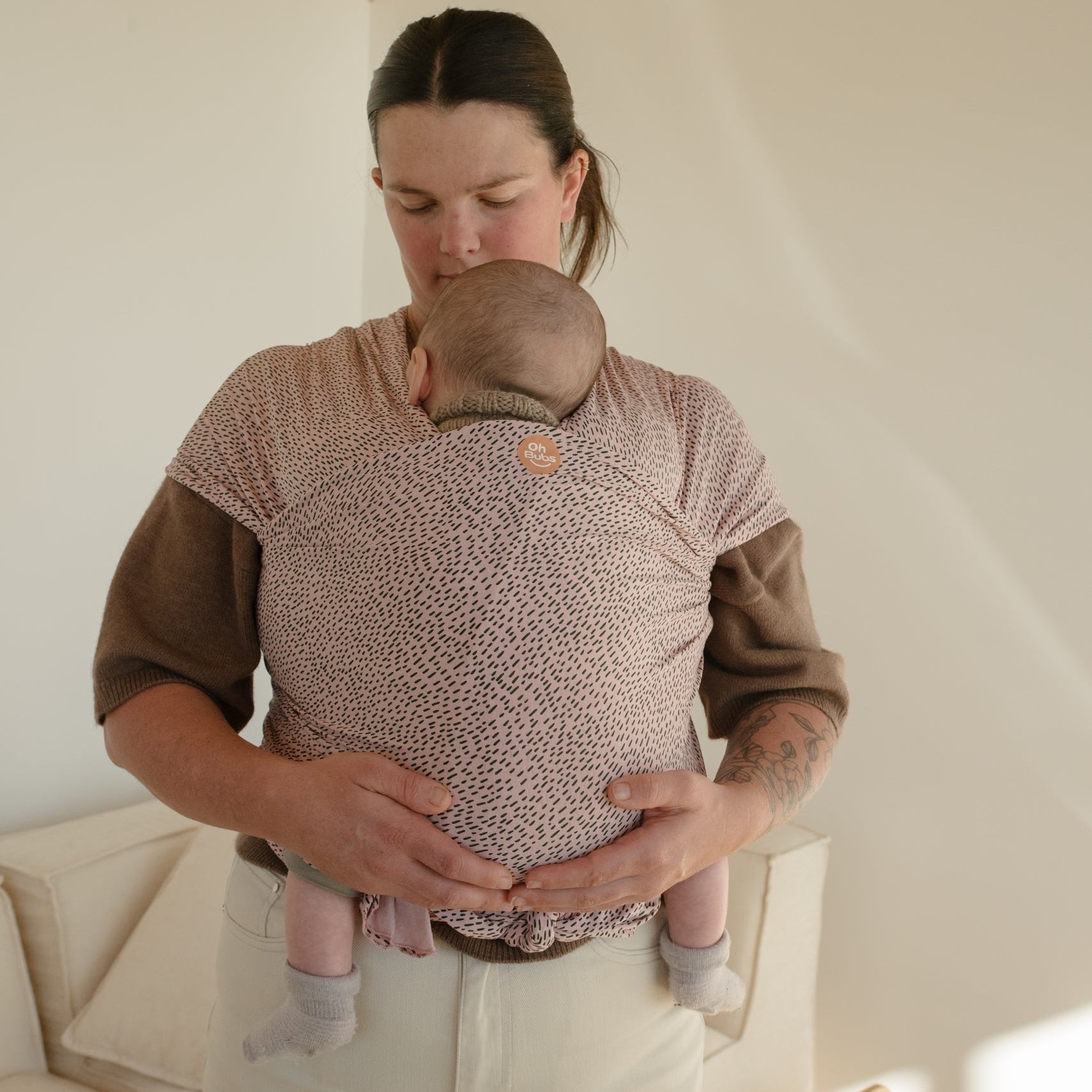 A woman wearing a baby carrier wrap with a small pink and brown pattern, holding a baby against her chest in a bright, modern room.