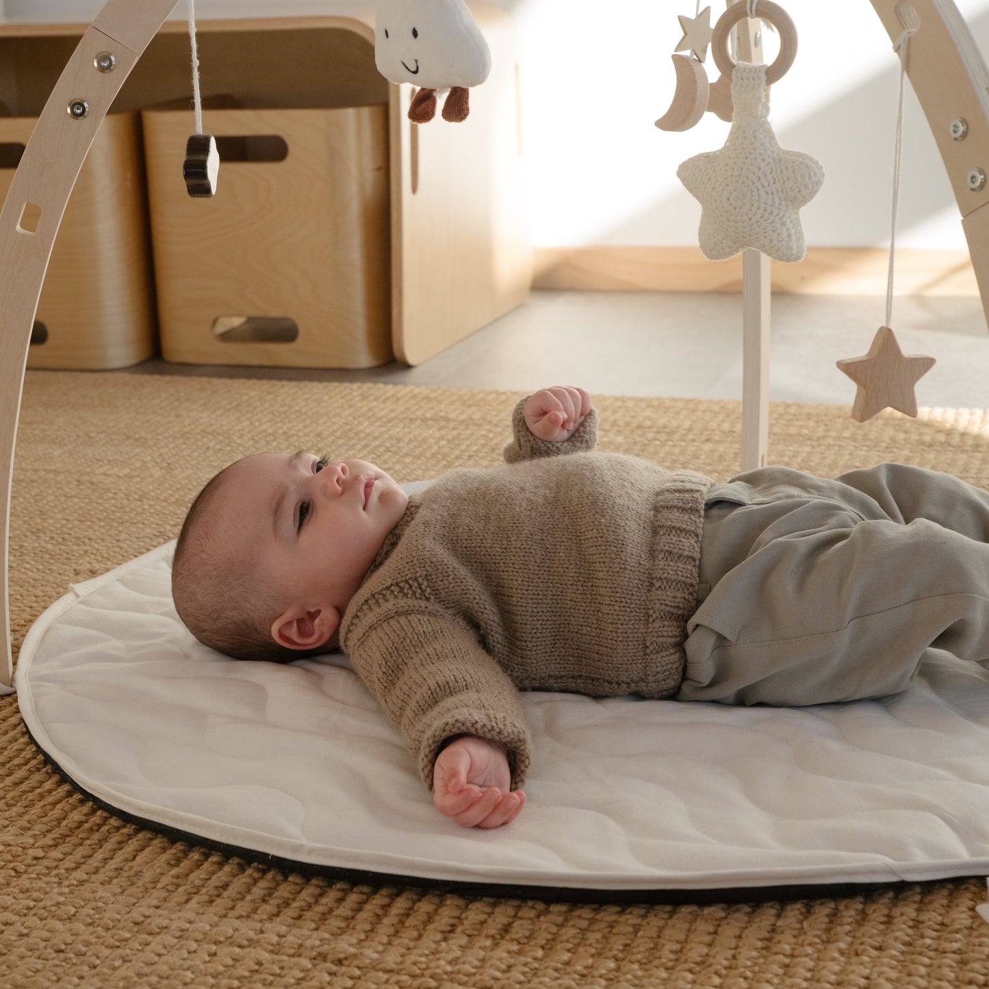 A baby lies on a round baby gym mat on a woven rug, wearing a beige knit sweater and gray pants. The baby gym has a light wood frame with hanging toys, including a star and a bunny, in a bright, modern nursery setting.
