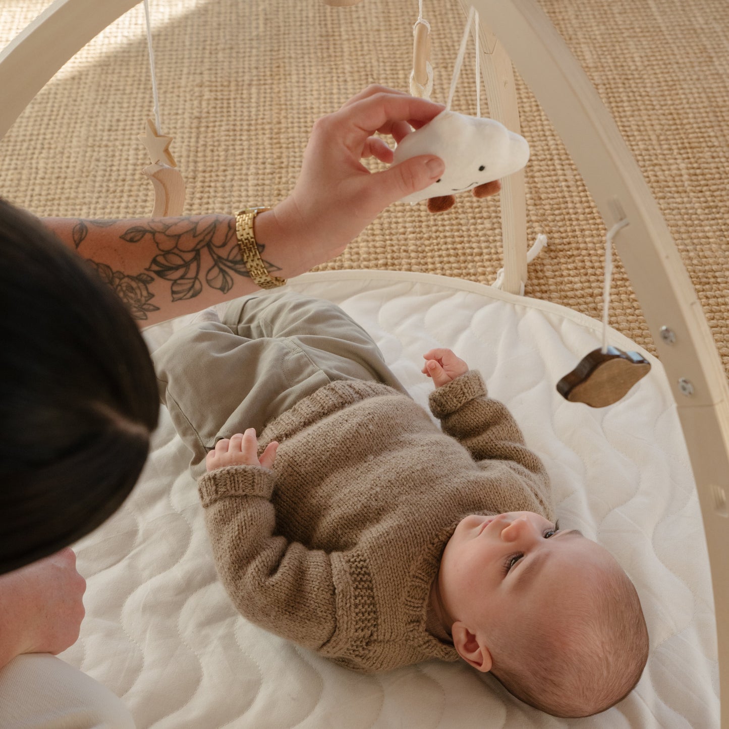 A baby in a beige knit outfit lies on a white padded baby gym mat while a person holds a white hanging toy. The scene is photographed in a warm, natural setting with a woven rug background.