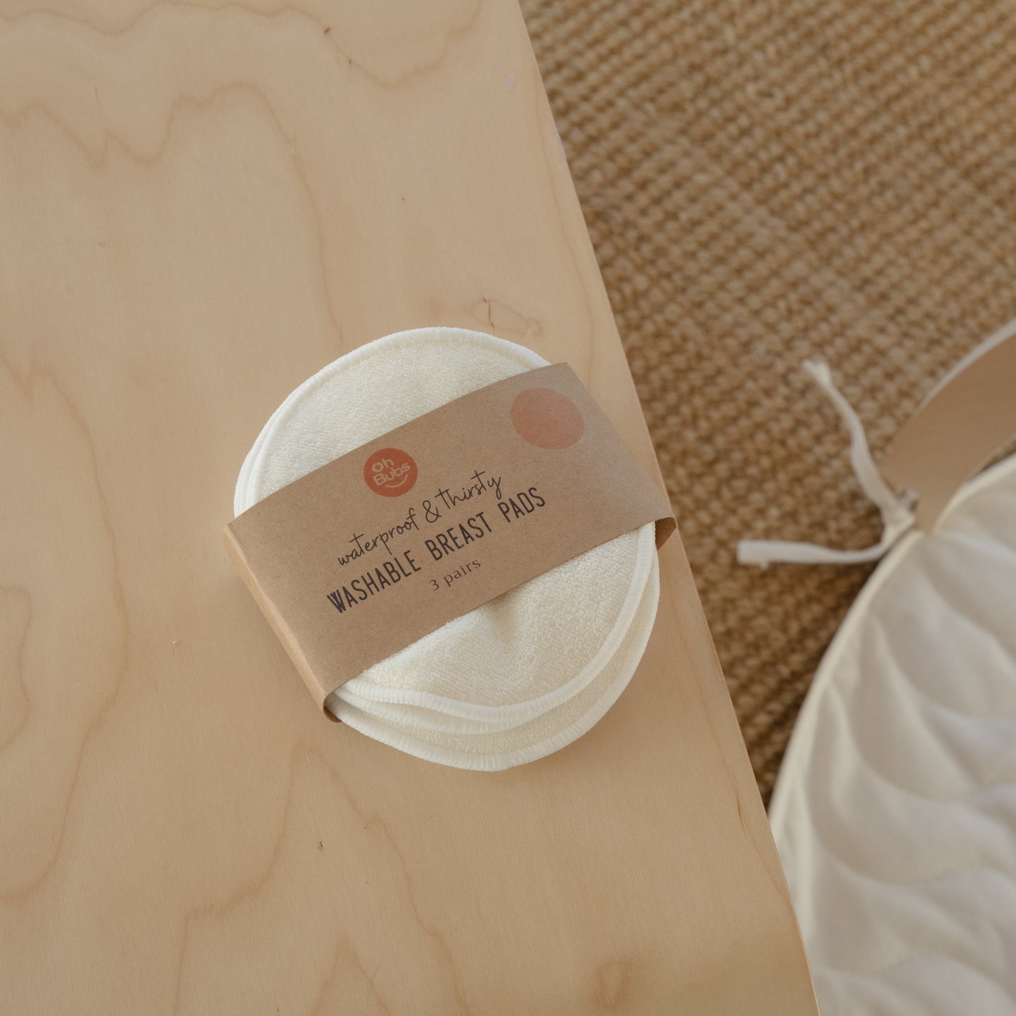 A small round container of washable breast pads wrapped in a kraft paper band, placed on a light wood surface with a white fabric bag nearby on a woven rug.