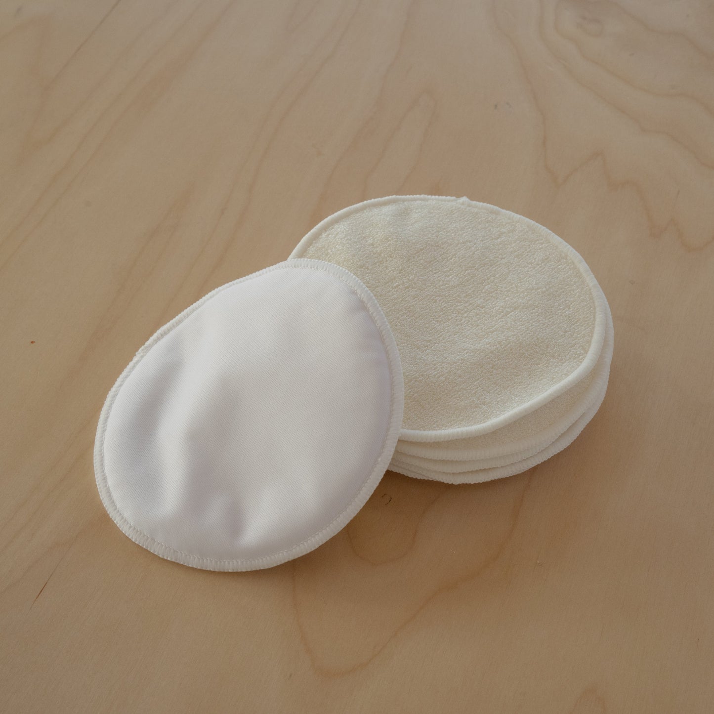 A stack of white reusable round breast pads resting on a light wood grain surface.